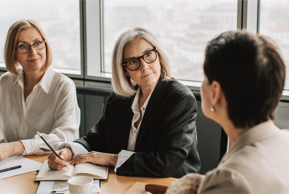 Two women with glasses listen to a third as she speaks.