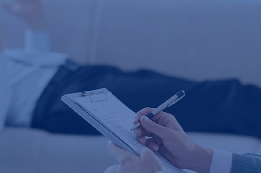Patient lies on couch in background while therapist takes notes.