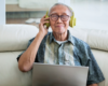 Stock image of an older Asian man sitting on a white couch and looking happy while wearing yellow headphones and listening to music on his computer.