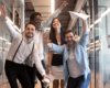 Stock image showing a diverse group of employees smiling and posing in celebration in a glass corridor of an office building.