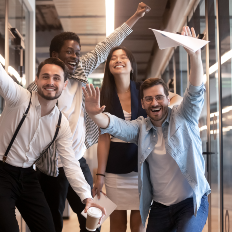 Stock image showing a diverse group of employees smiling and posing in celebration in a glass corridor of an office building.