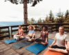 Stock image of four women sitting in lotus position on yoga mats outside on a porch with a tree in the background.