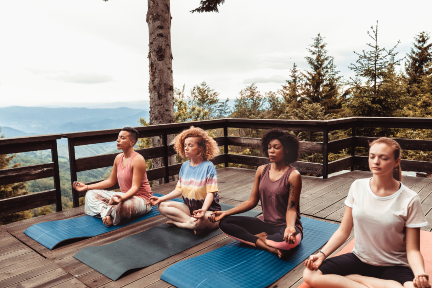 Stock image of four women sitting in lotus position on yoga mats outside on a porch with a tree in the background.