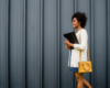 Stock image of professional black woman walking past a black wall wearing a blue dress, white cardigan, yellow purse and carrying a folder.