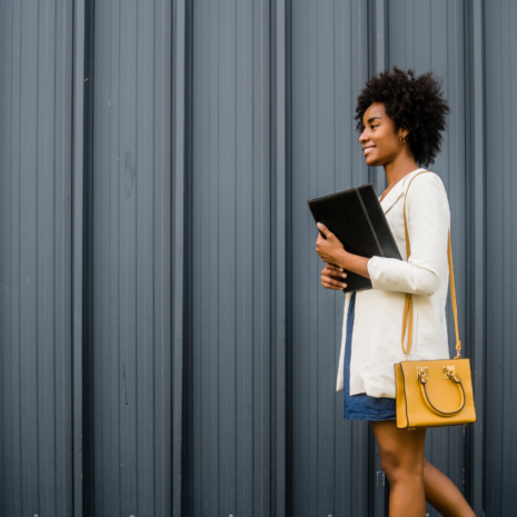 Stock image of professional black woman walking past a black wall wearing a blue dress, white cardigan, yellow purse and carrying a folder.