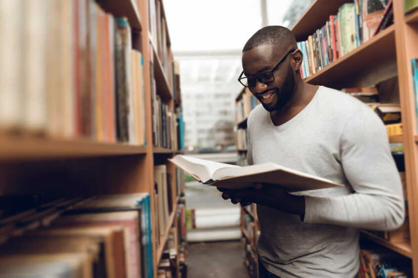 Ethnic African American Guy Reading Book, Smiling In Aisle Of Library