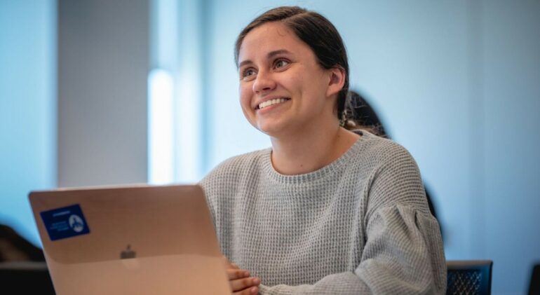 A young woman seating in front of a laptop computer smiles and looks off-camera.