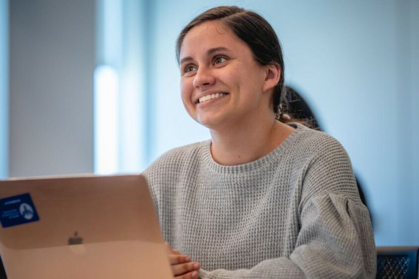 A young woman seating in front of a laptop computer smiles and looks off-camera.