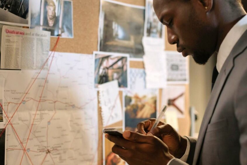 A criminologist studies his notes while standing in front of a bulletin board covered with documents.