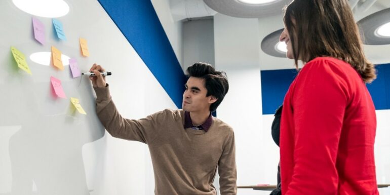 A man writes on a whiteboard covered with sticky notes while a woman looks on.