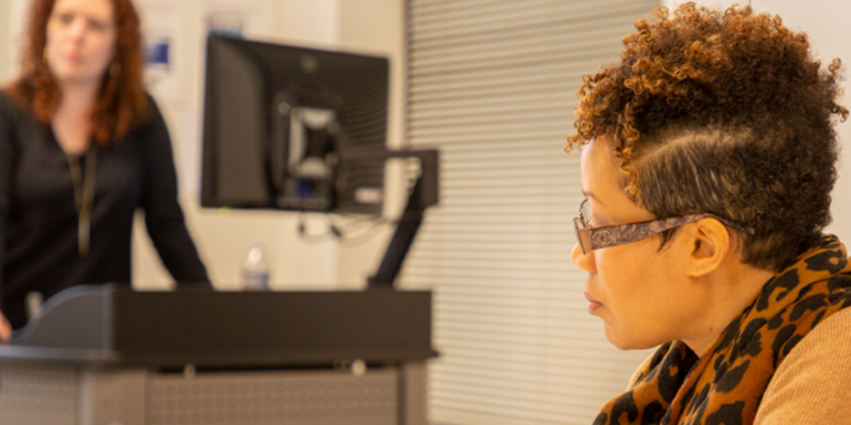 A woman sits in the foreground while a woman standing in the background gives a presentation.