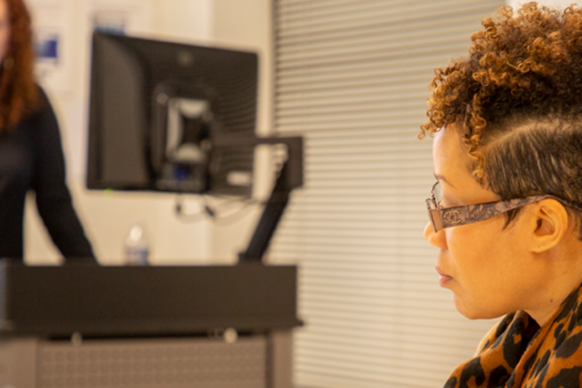 A woman sits in the foreground while a woman standing in the background gives a presentation.