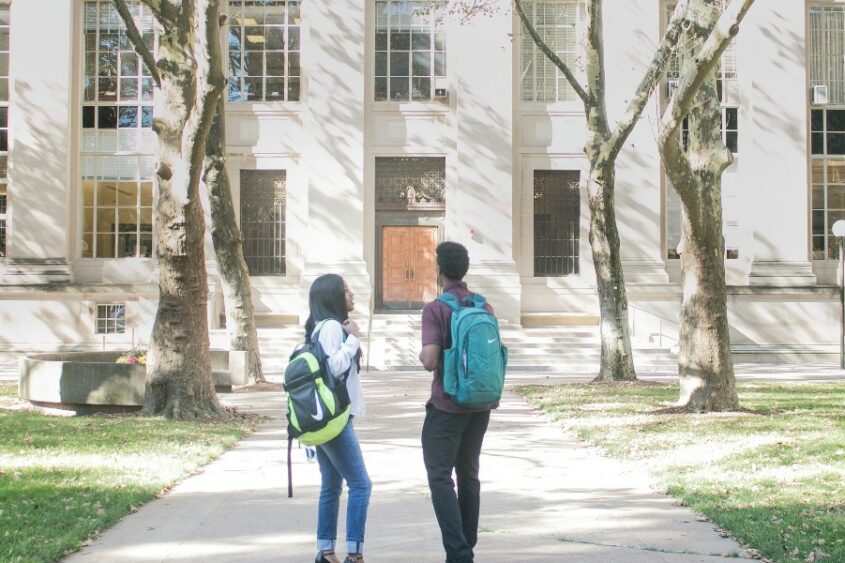 two students wearing backpacks outside a stone college building