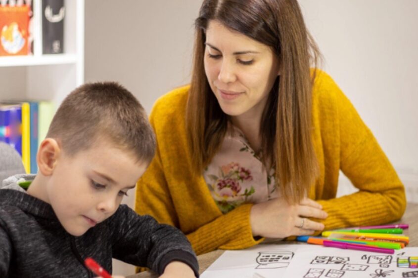 A young woman looks on in a classroom as a little boy draws with a red marker.
