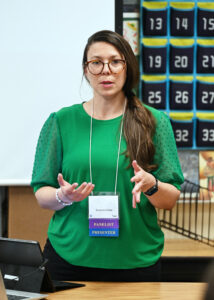 Woman with glasses and green top standing and speaking in a classroom.