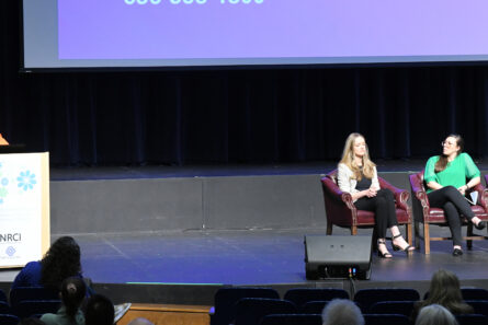 Three women sitting on stage watching a fourth speaking.