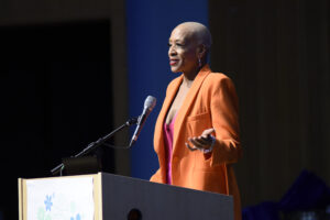 African American woman with short hair and orange jacket speaking from a podium.