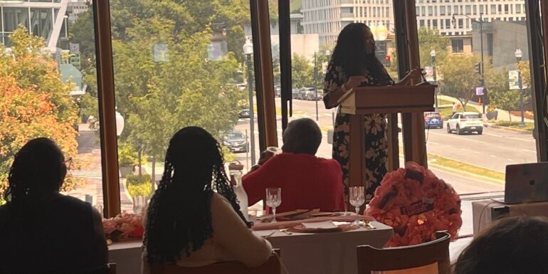 A woman speaks at a podium next to a window.