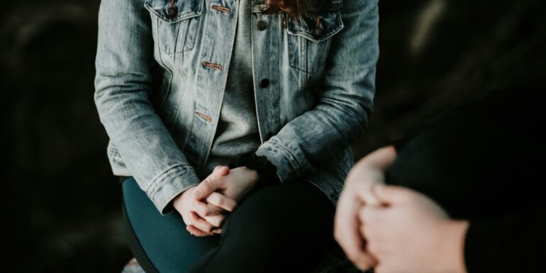 A close-up of two people's folded hands as they sit next to each other