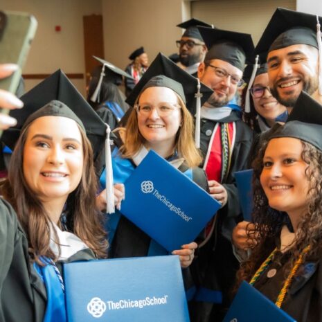 Newly graduated students from The Chicago School, wearing caps and gowns, pose for a group selfie holding their freshly issued diplomas.