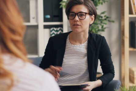 A woman with glasses talking with another woman mostly out of frame.