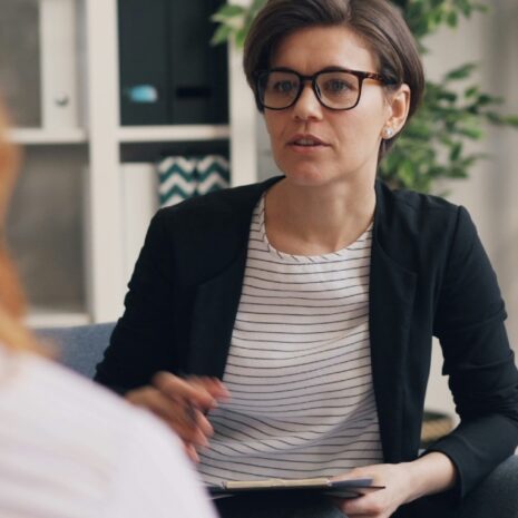 A woman with glasses talking with another woman mostly out of frame.
