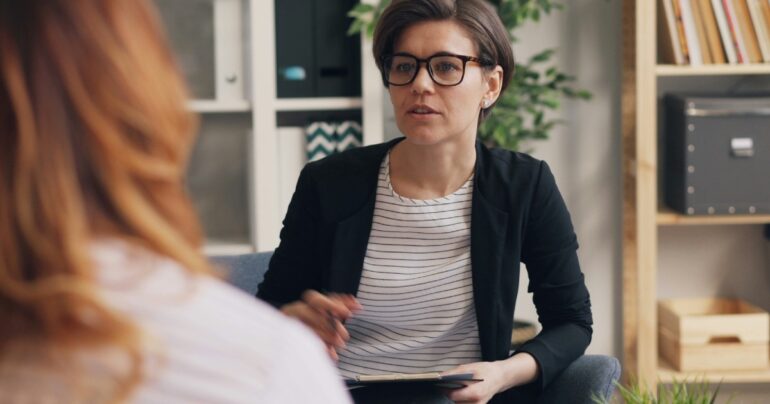 A woman with glasses talking with another woman mostly out of frame.