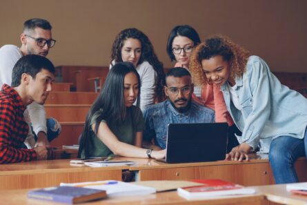 Adult students in a classroom gather around a laptop.
