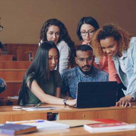Adult students in a classroom gather around a laptop.