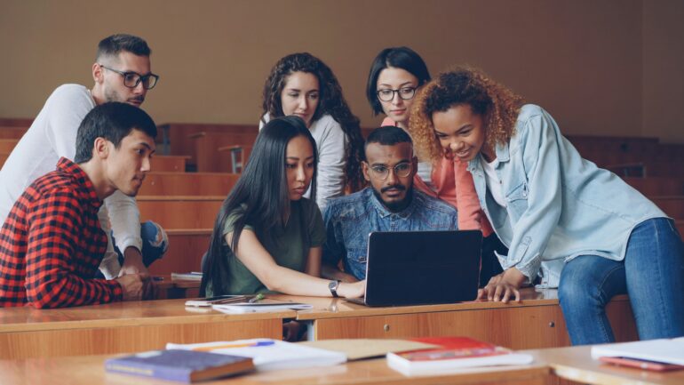 Adult students in a classroom gather around a laptop.