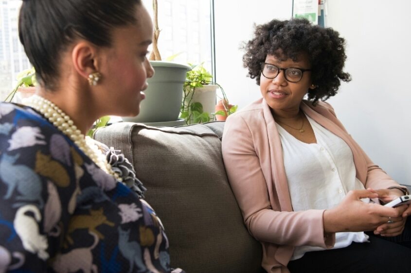 A woman sitting on a couch in a sunlit room speaks to another woman sitting next to her on the ouch.