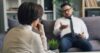 Man on a couch talks to a woman in the foreground holding a pen.