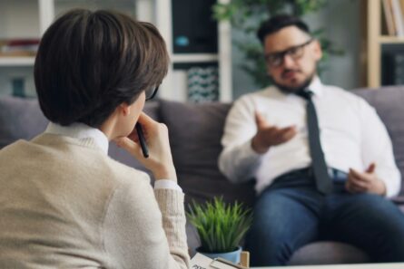 Man on a couch talks to a woman in the foreground holding a pen.