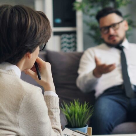 Man on a couch talks to a woman in the foreground holding a pen.