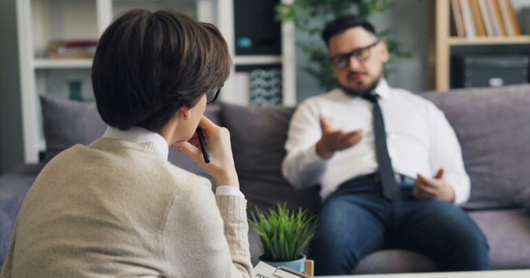 Man on a couch talks to a woman in the foreground holding a pen.
