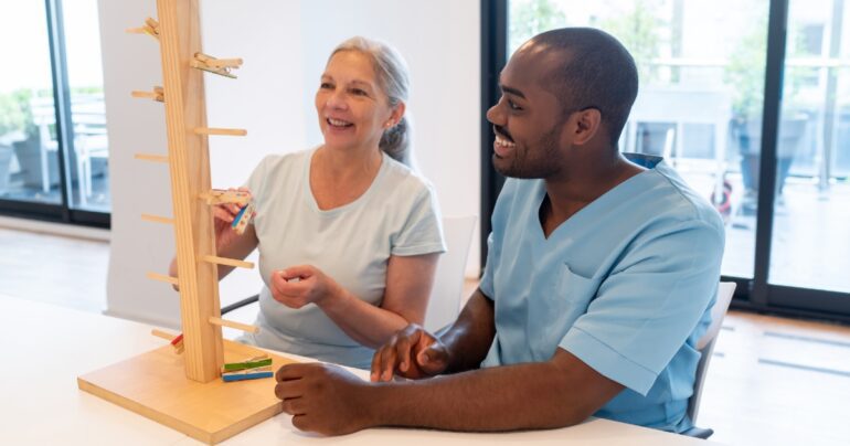 Older woman works with an occupational therapist on a wooden clothespin activity to practice fine motor skills and hand coordination.