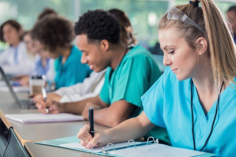 Diverse male and female young medical students confidently take notes during their professor's lecture. They are wearing scrubs.