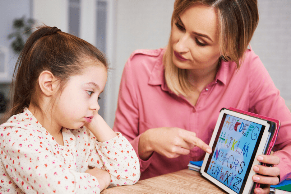 woman in pink shirt pointing to letters on a tablet with young child